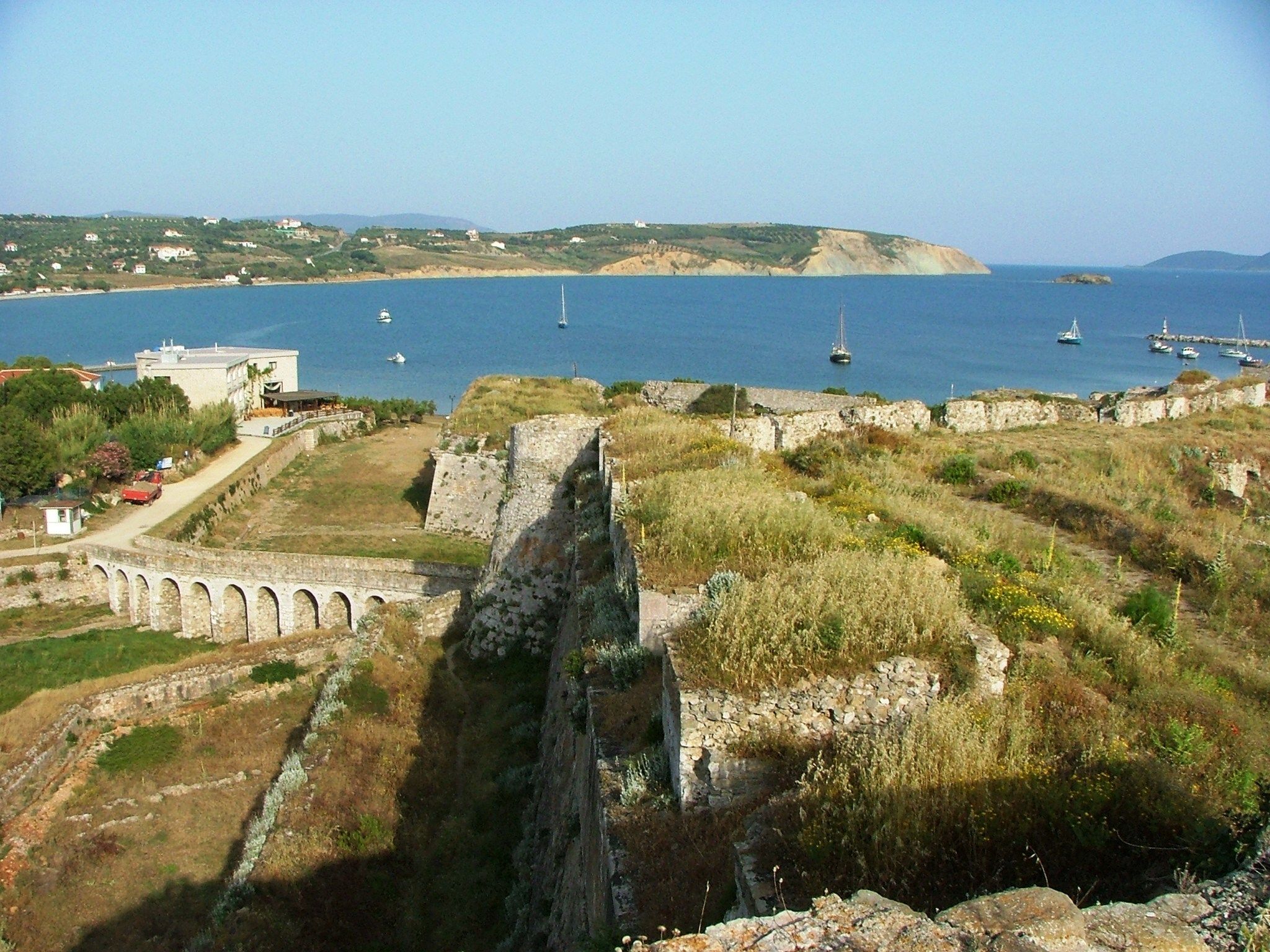 Un sitio histórico costero con ruinas, un puente y barcos al fondo.