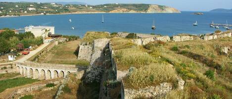 Un sitio histórico costero con ruinas, un puente y barcos al fondo.