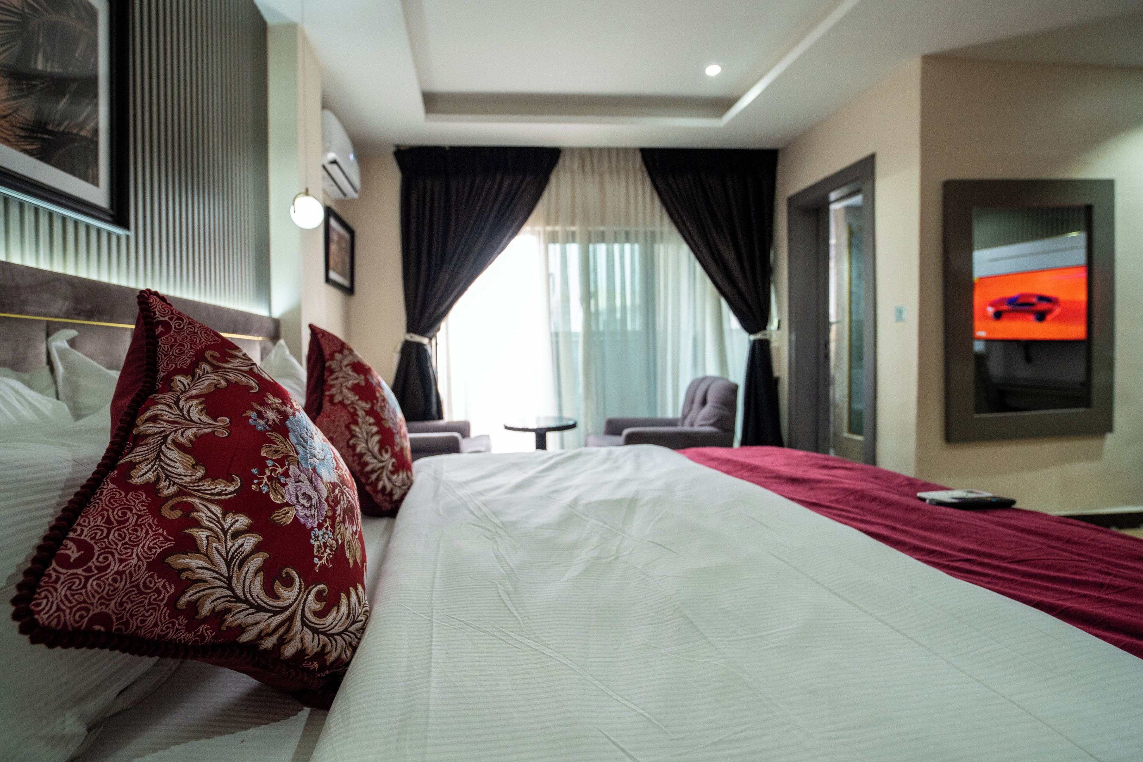 Quarto de hotel com uma cama grande, duas almofadas decorativas vermelhas, uma televisão na parede e um cobertor vermelho.
