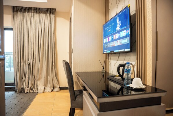 A hotel room with a bed, a desk with a computer and a monitor, a chair, a window with curtains, and a water bottle.