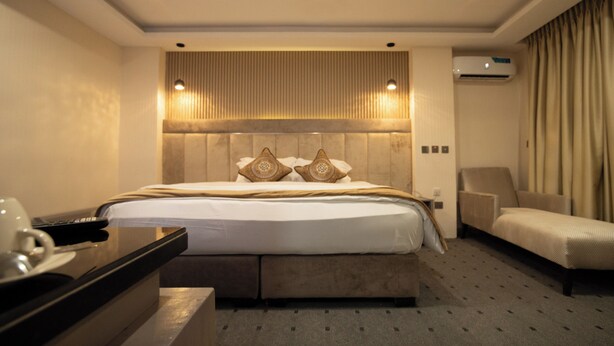 A hotel room with a large bed, a bedside table, a chair, and an air conditioning unit. - Vynedresa Hotel (Abuja)