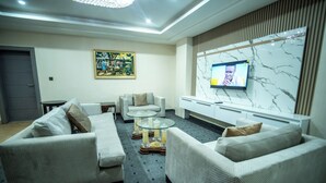 A modern living room with a flat-screen TV mounted on a marble wall, two sofas, a glass coffee table, and a small side table. - Vynedresa Hotel (Abuja)