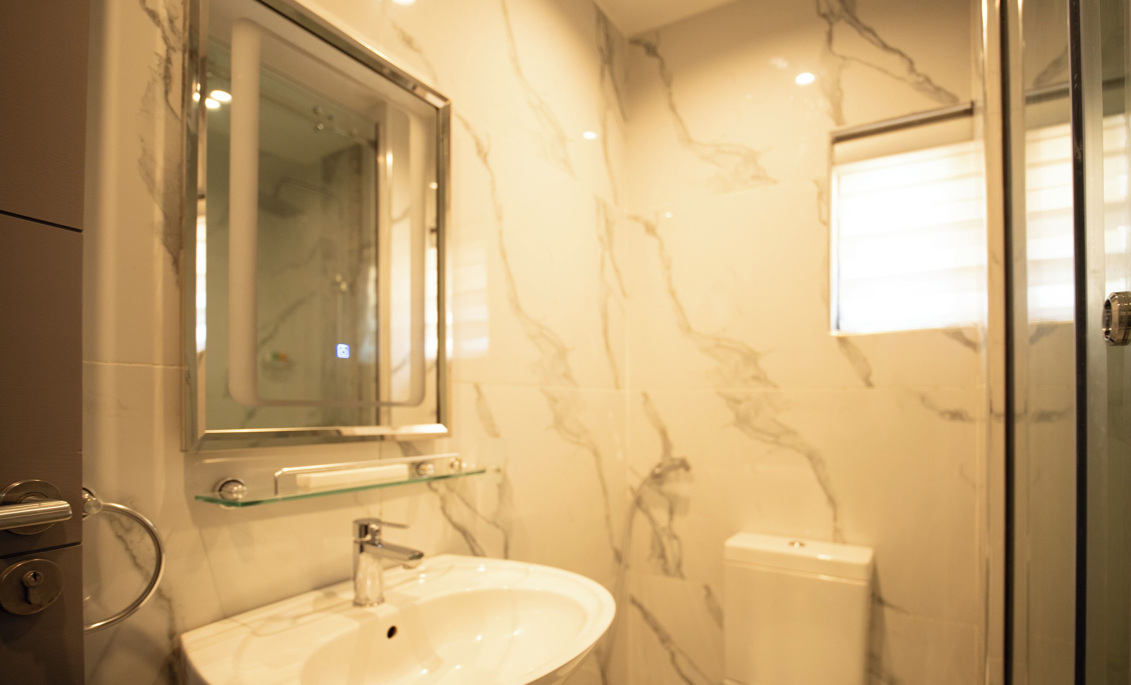 A bathroom with a marble wall, a glass shelf above a sink, and a toilet.