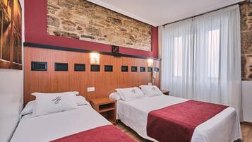 Une chambre d’hôtel avec deux lits, un mur en pierre, une tête de lit en bois et une grande fenêtre avec des rideaux blancs.