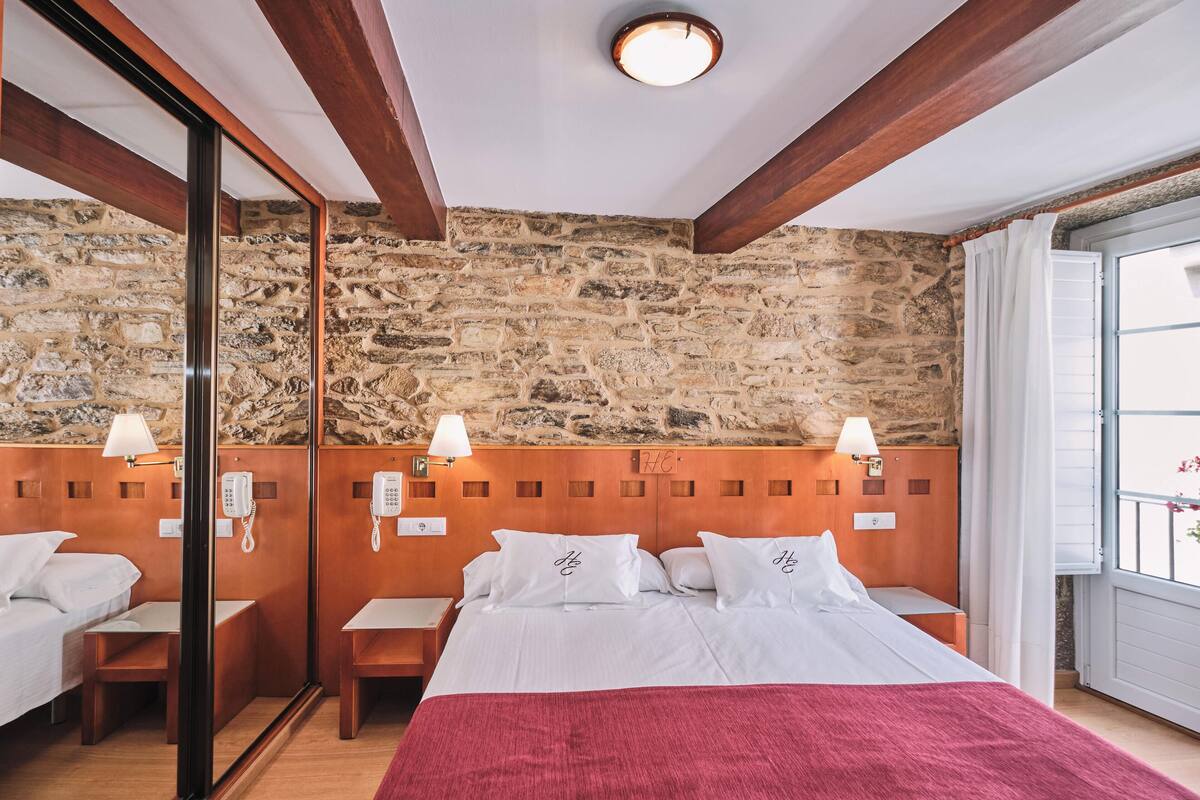 Une chambre d’hôtel avec un mur en pierre, une tête de lit en bois, un lit avec une literie blanche, des tables de chevet, des lampes et une fenêtre avec des rideaux.