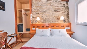 Quarto de hotel com parede de pedra, cabeceira de madeira, duas luminárias de apoio, cama com roupa de cama branca, uma cadeira de madeira e uma escrivaninha.