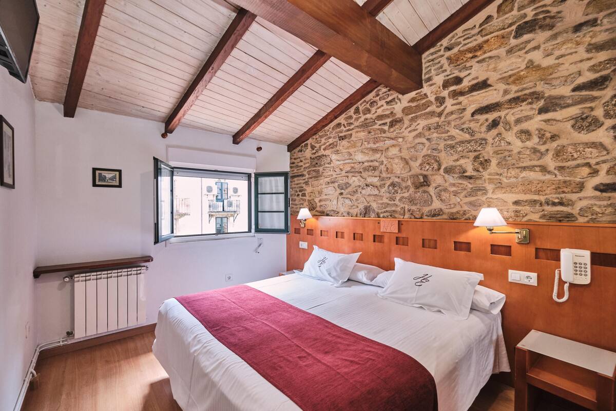 Une chambre avec un mur en pierre, des poutres en bois, un lit avec du linge de lit blanc et une couverture rouge, une table de chevet, une lampe et un téléphone.
