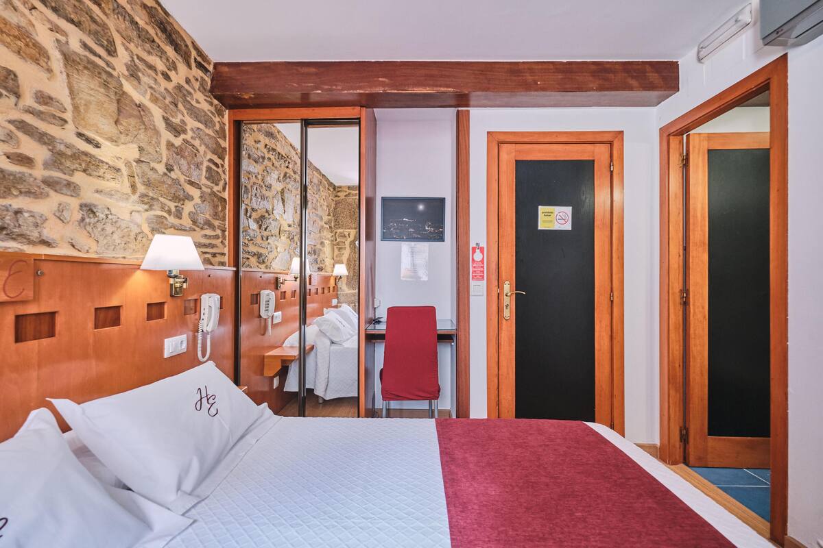 Une chambre d’hôtel avec un mur en pierre, une tête de lit en bois, un lit avec du linge de maison blanc, un bureau avec une chaise et une porte avec une pancarte.