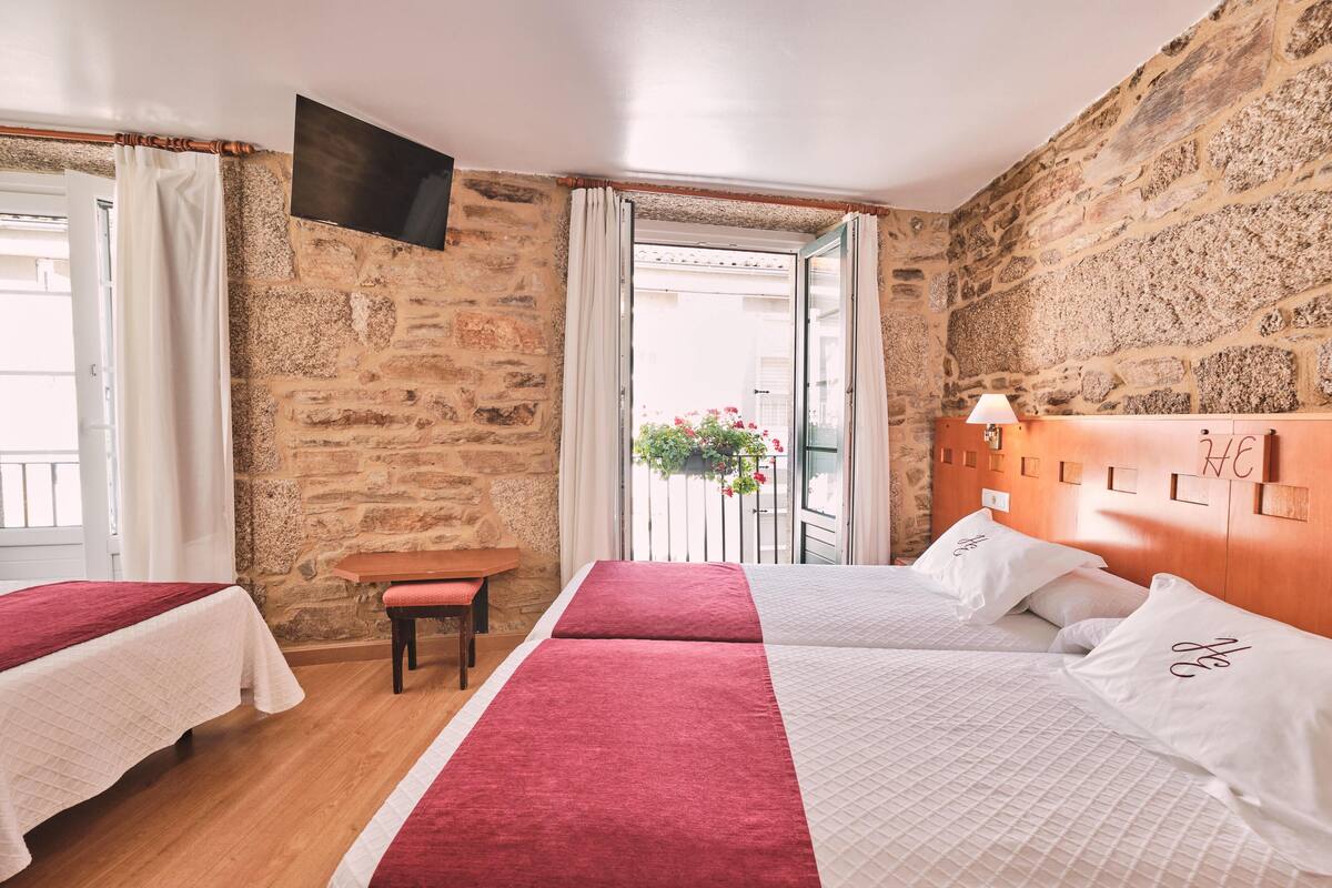 Une chambre d’hôtel avec deux lits, une tête de lit en bois, une petite table, un banc et un mur en pierre.