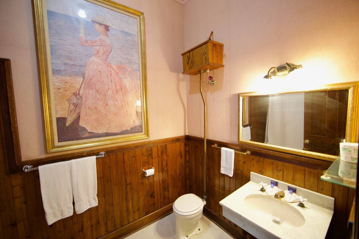 A bathroom with wooden paneling, a large framed painting, a toilet, a sink, and a mirror.