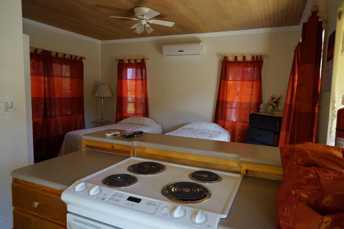 A compact room with a kitchenette, two beds, a ceiling fan, and red curtains.