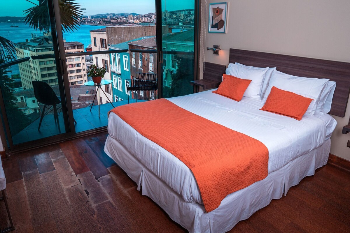 Ein Hotelzimmer mit einem großen Bett, einem Balkon mit Blick auf die Stadt und einem kleinen Tisch mit einem Stuhl.