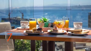 Una mesa de desayuno con jugo de naranja, un muffin, tostadas y un bol de fruta, con vista a un paisaje panorámico de una masa de agua y un horizonte urbano distante.