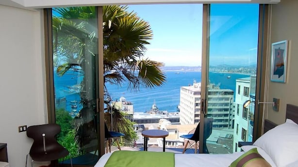 Habitación de hotel con un ventanal que ofrece vistas al mar y al paisaje urbano.