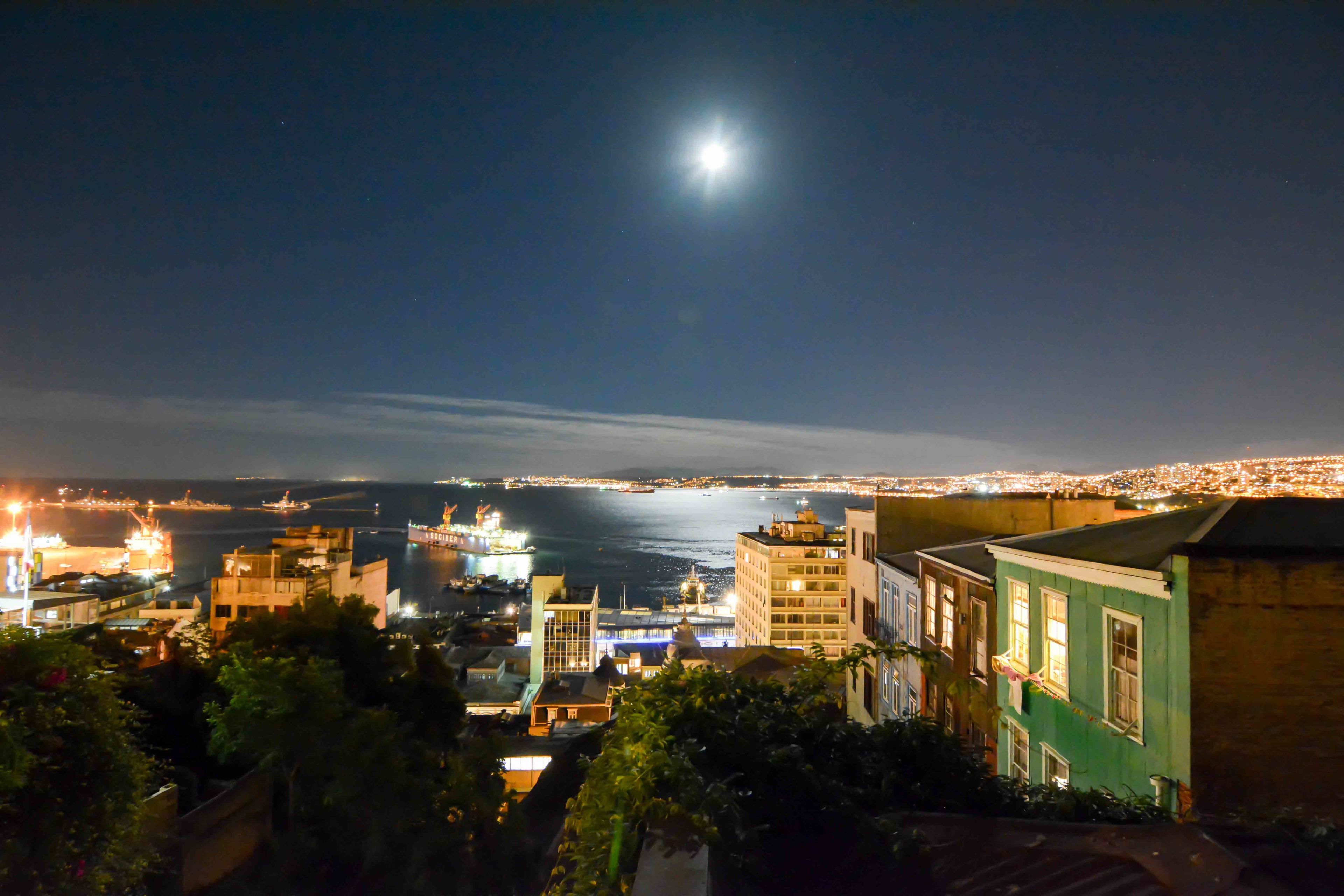 A nighttime cityscape with a prominent moon, buildings, and a body of water.