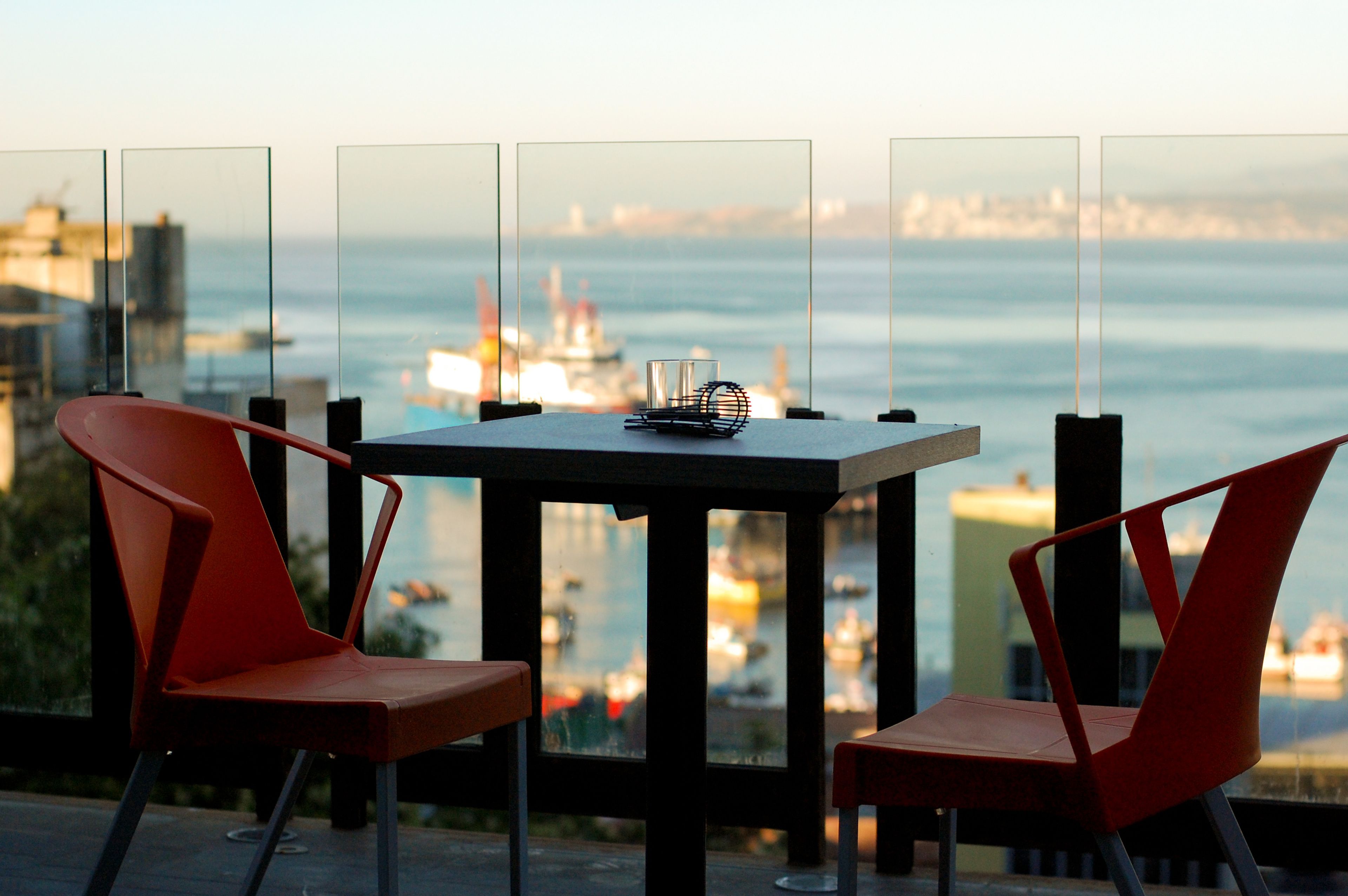 Ein Tisch mit zwei roten Stühlen, einem Glas und einem Metallgegenstand, vor einer Aussicht auf das Meer und Gebäude.