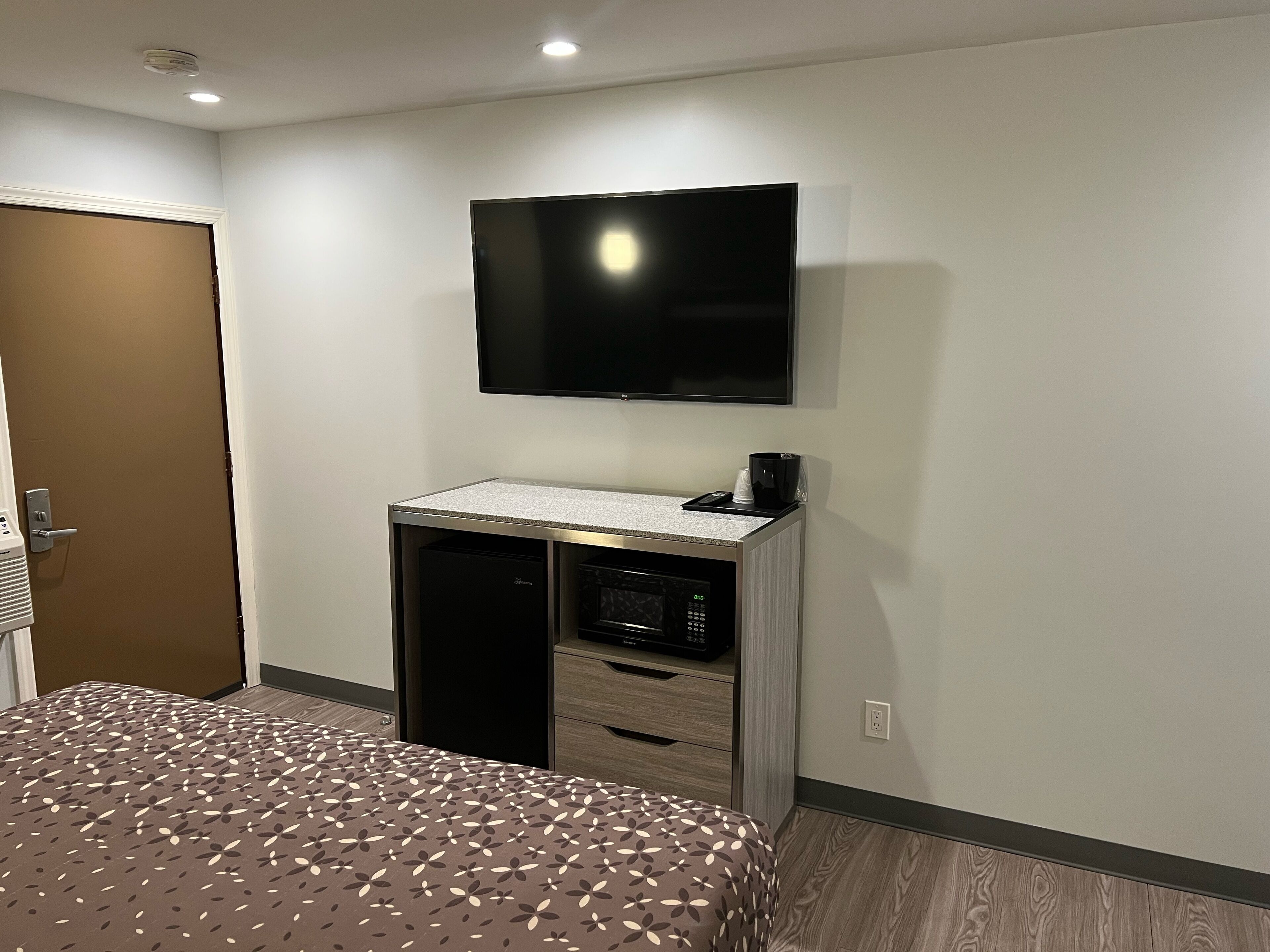 A hotel room with a bed, a TV mounted on the wall, a minibar, and a door.