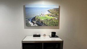 Quarto de hotel com uma grande pintura de paisagem costeira, uma escrivaninha com um expositor de lenços, um relógio e uma cafeteira.