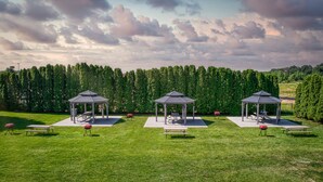 Trois kiosques abritant des tables de pique-nique et des bancs, situés dans une zone herbeuse, avec une haie dense en arrière-plan.
