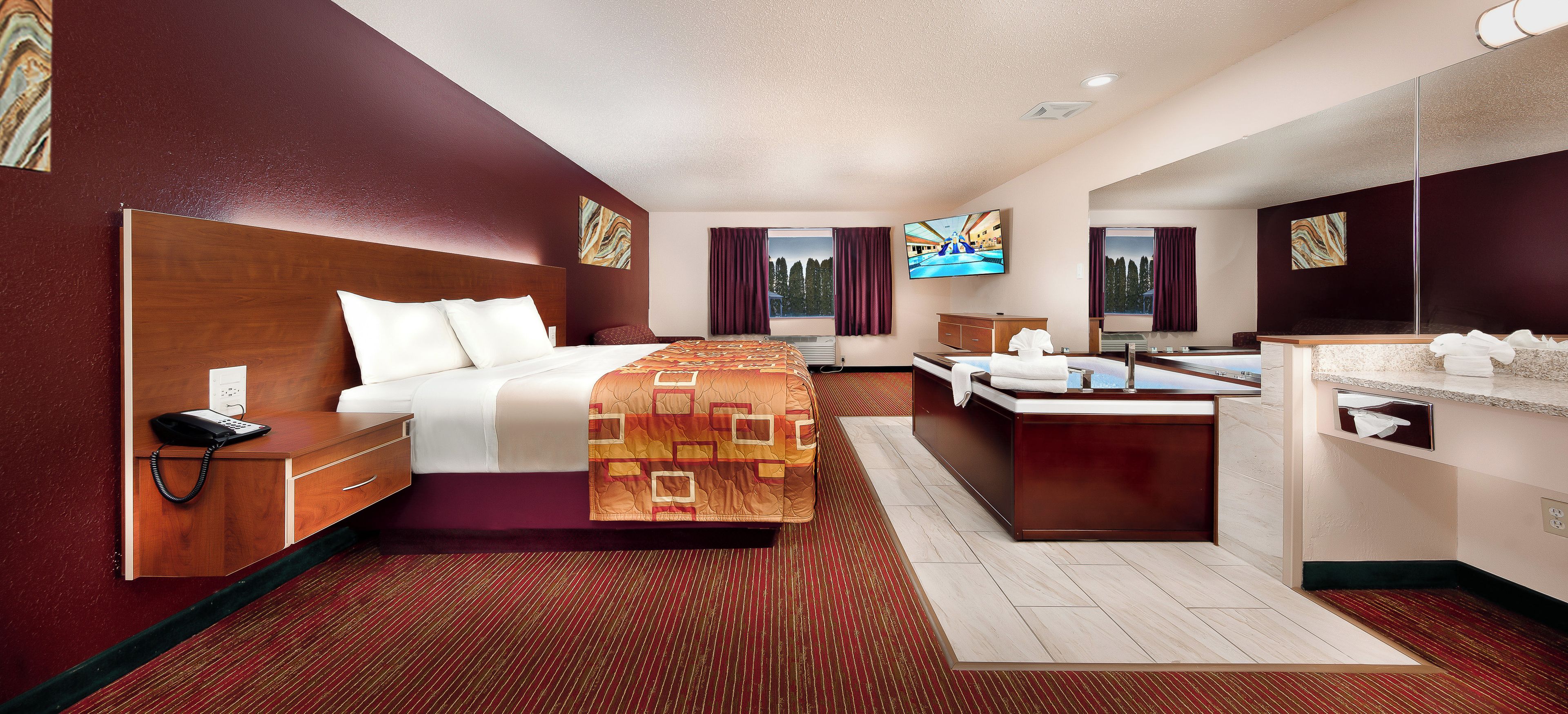 A hotel room with a bed, bedside table with a phone, a desk with a flat-screen TV, a bathroom with a sink and mirror, and a large window with curtains.