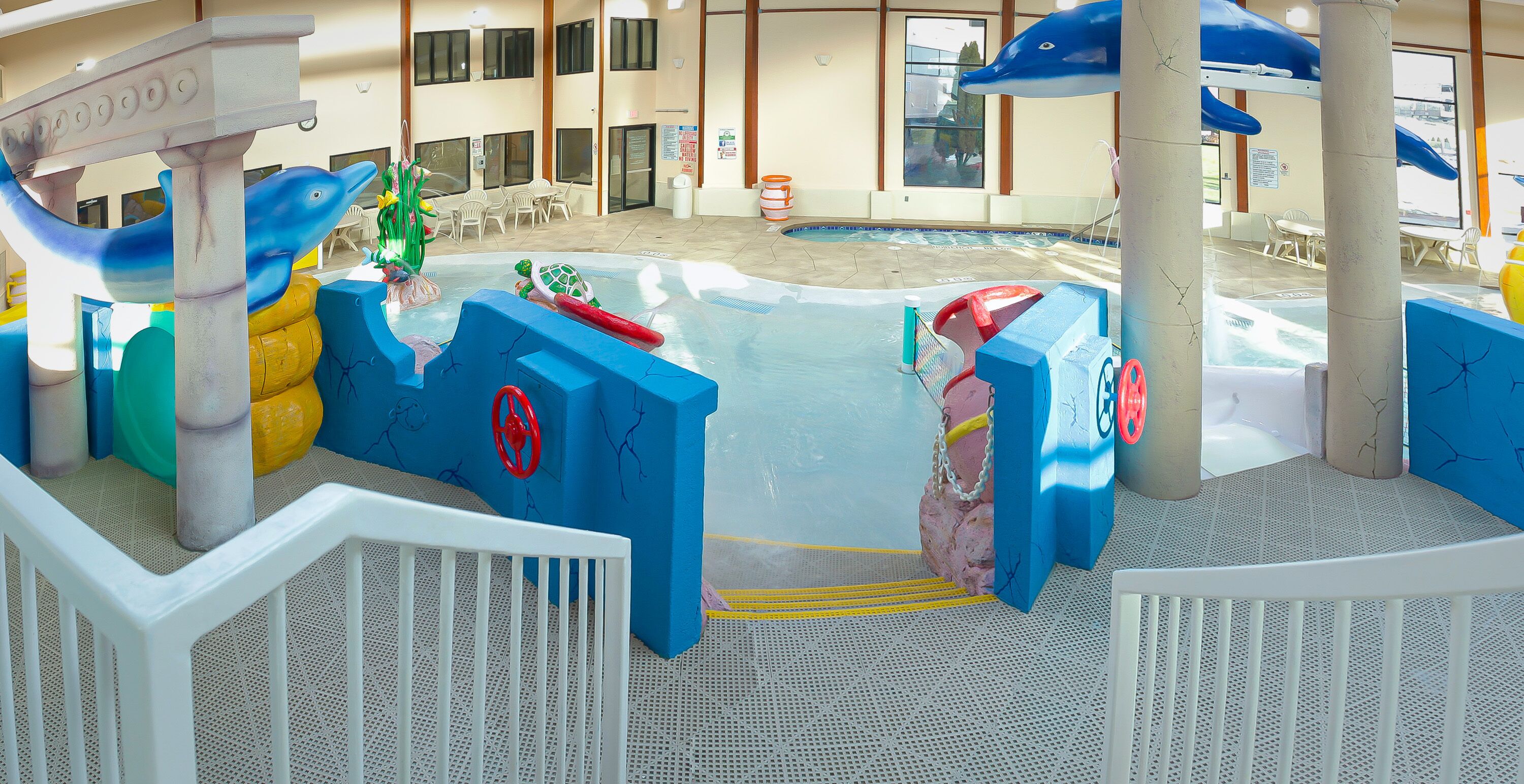 A water play area with slides and a pool, surrounded by a white fence and a building with large windows in the background.