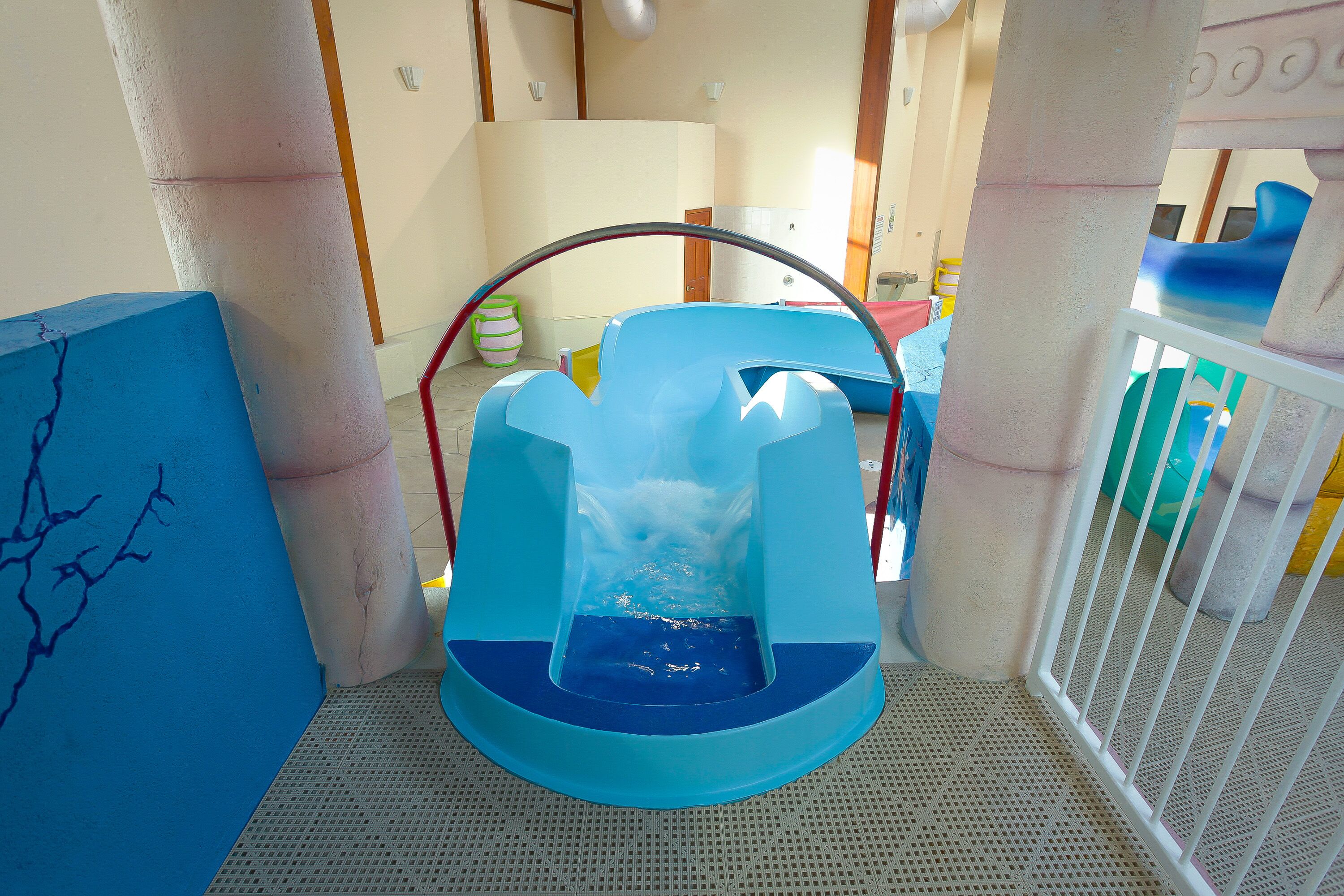 A blue slide with a water feature, located in a room with white pillars and a white railing.