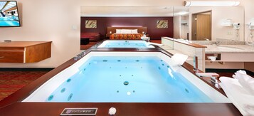 A hotel room with a large spa area, a bed, a TV, and a bathroom with a mirror and towel rack.