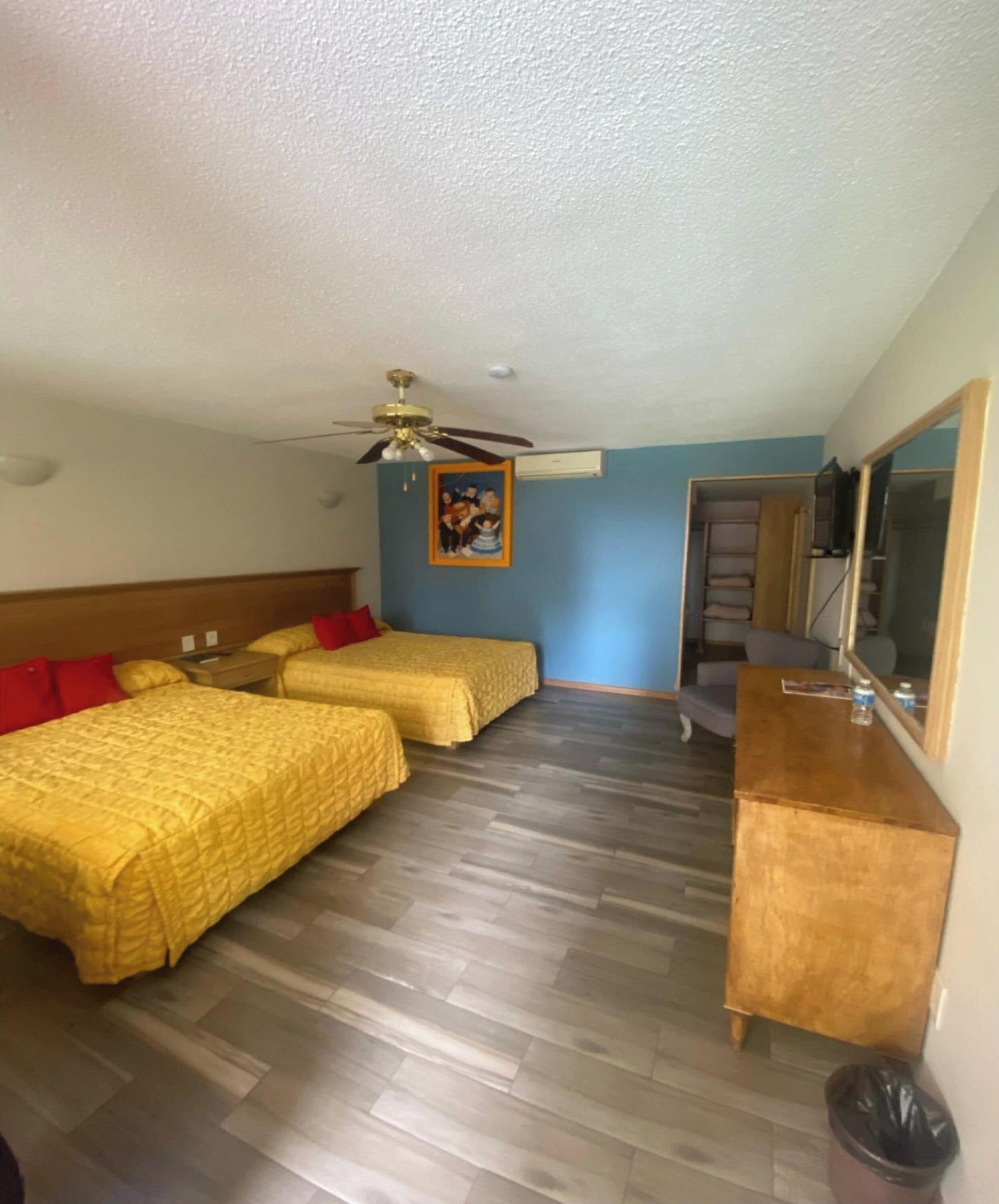A hotel room with two beds, a wooden desk, a ceiling fan, and a painting on the wall.