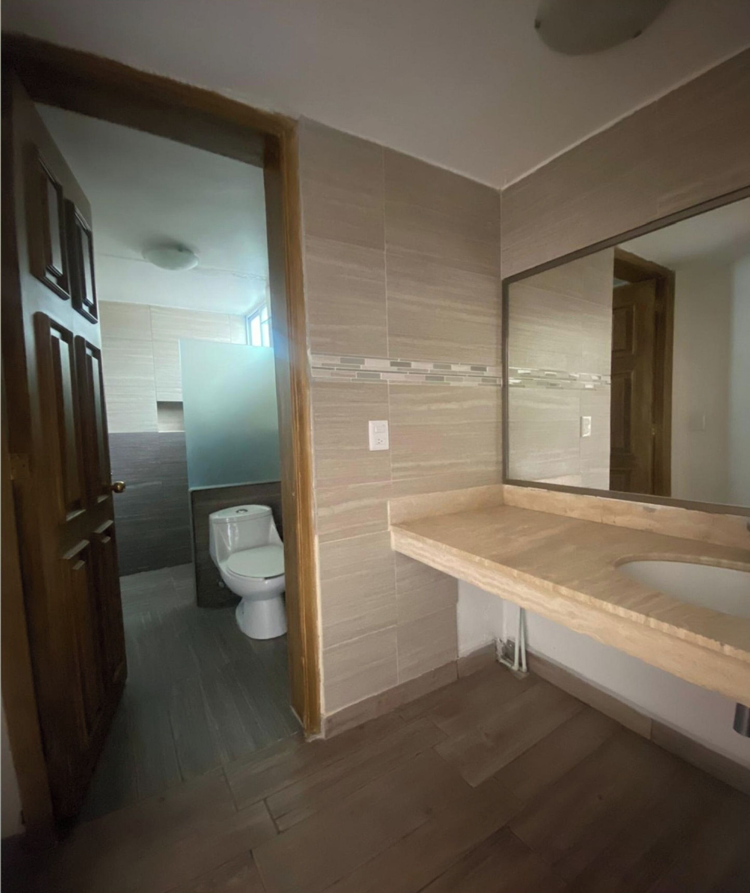 Un baño con puerta de madera, un inodoro, un lavabo con espejo y paredes con azulejos.