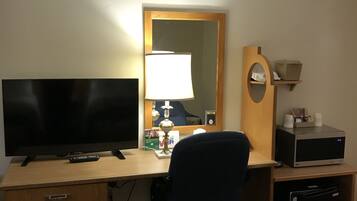 Ein Hotelzimmer mit einem Holztisch, einem Fernseher, einer Lampe, einer Mikrowelle und einem kleinen Regal mit Gegenständen.