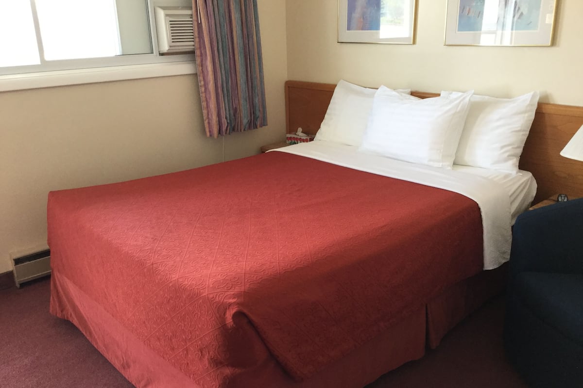 A hotel room with a large bed, a red bedspread, white pillows, a wooden headboard, a window with curtains, and a small bedside table.