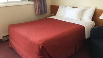 Camera d'albergo con un letto grande, una coperta rossa, cuscini bianchi, una testiera in legno, una finestra con tende e un comodino.