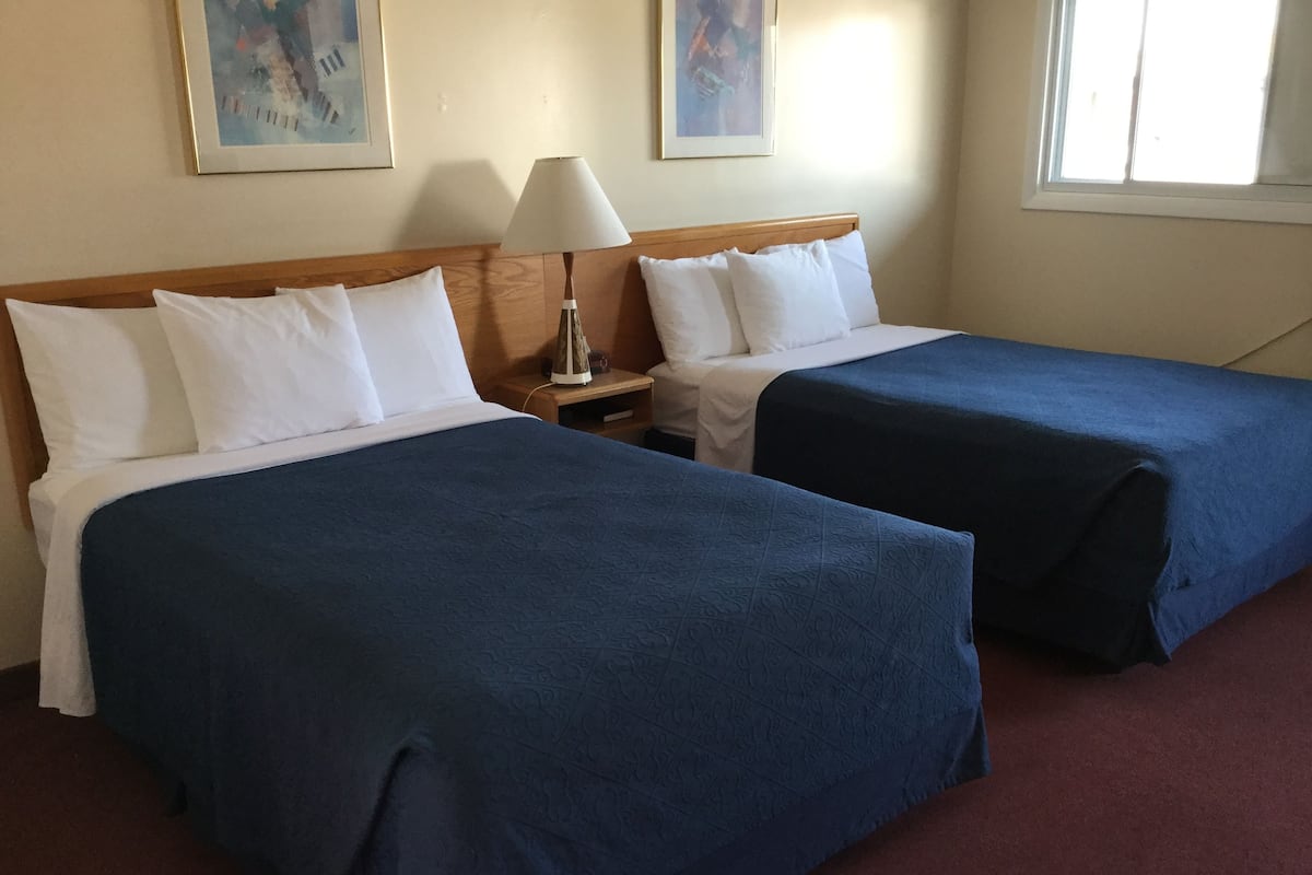 A hotel room with two beds, each with white pillows and blue bedspreads. There is a nightstand with a lamp and a framed picture on the wall.