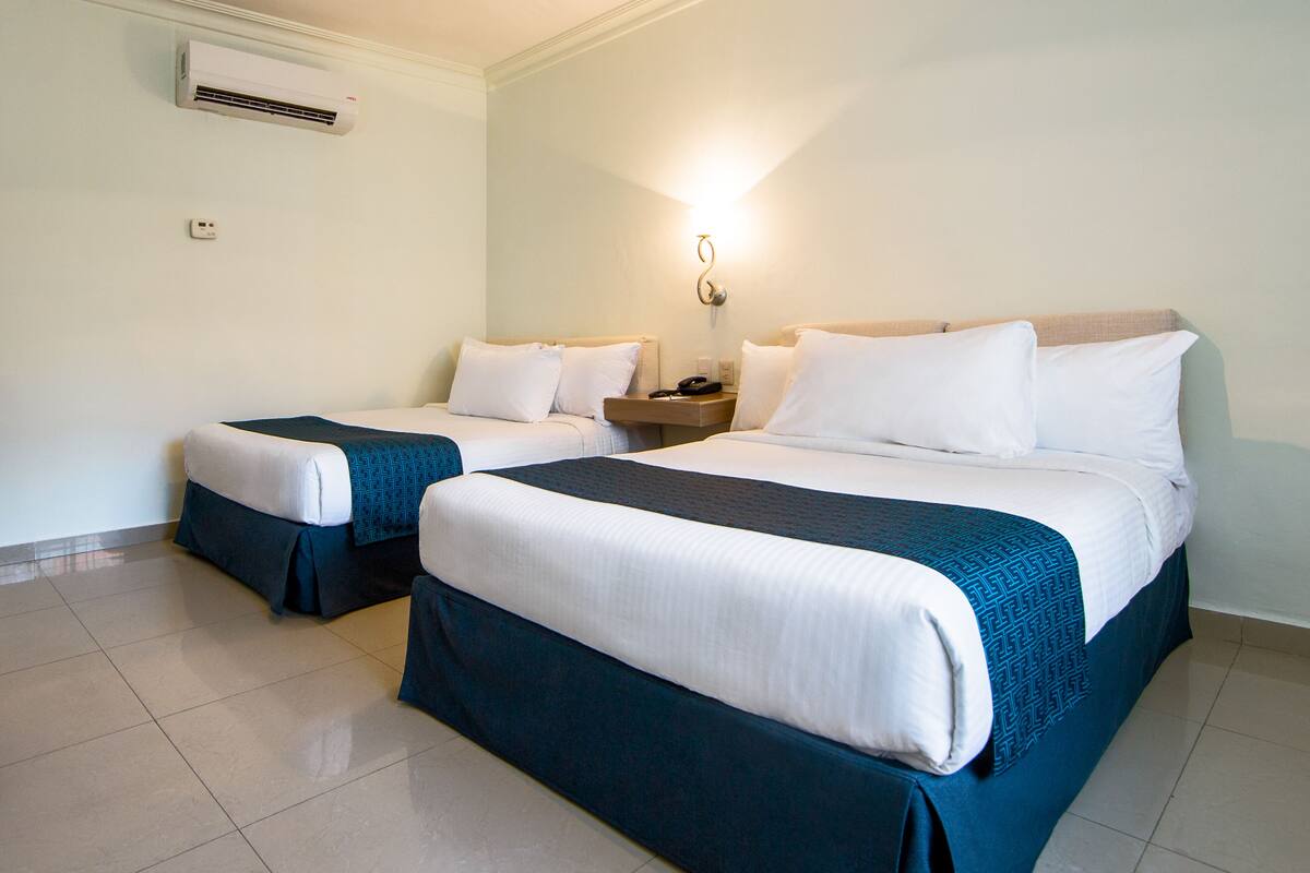 Habitación de hotel con dos camas, cada una con ropa blanca y cubrecamas color turquesa, una mesita de noche con lámpara y un equipo de aire acondicionado en la pared.