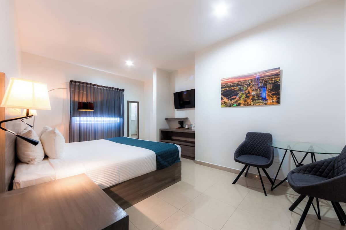 Habitación de hotel con cama, mesita de noche, lámpara, televisor, escritorio, sillas y un cuadro en la pared.