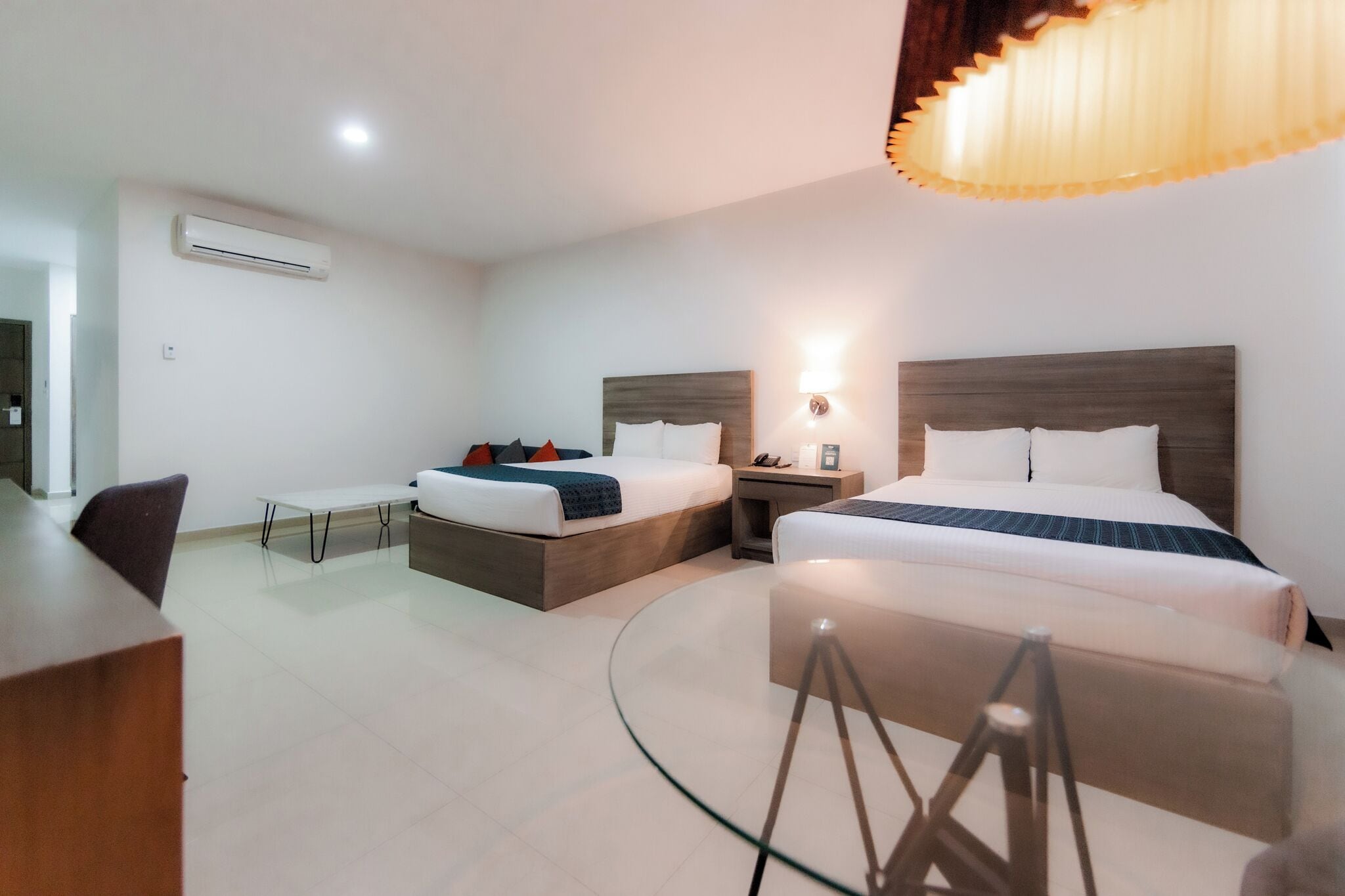 Una habitación de hotel moderna con dos camas, un escritorio, una mesa de vidrio y una lámpara en el techo.