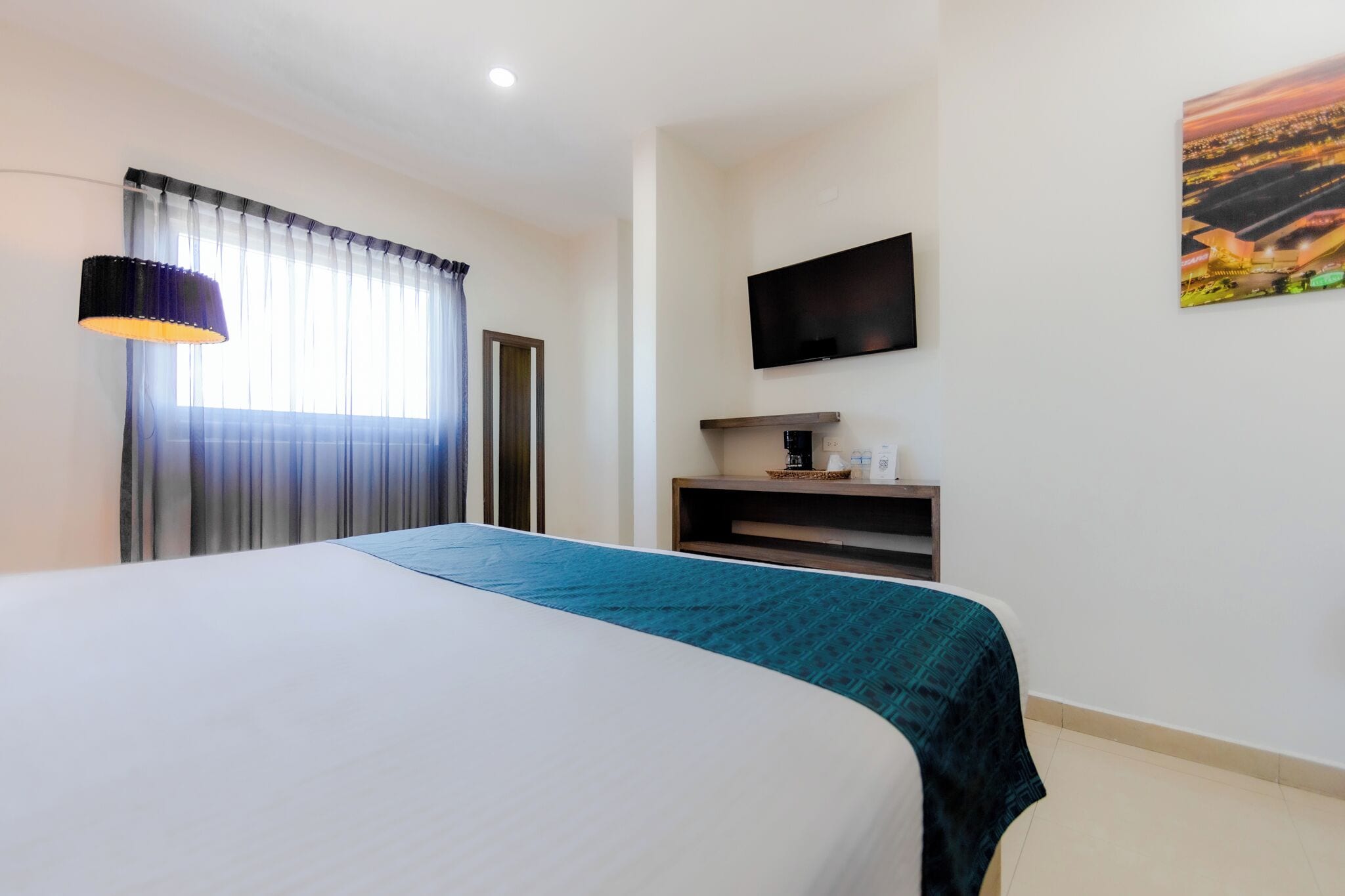 Habitación de hotel con una cama grande, un televisor de pantalla plana en la pared, una ventana con cortinas y un cuadro en la pared.