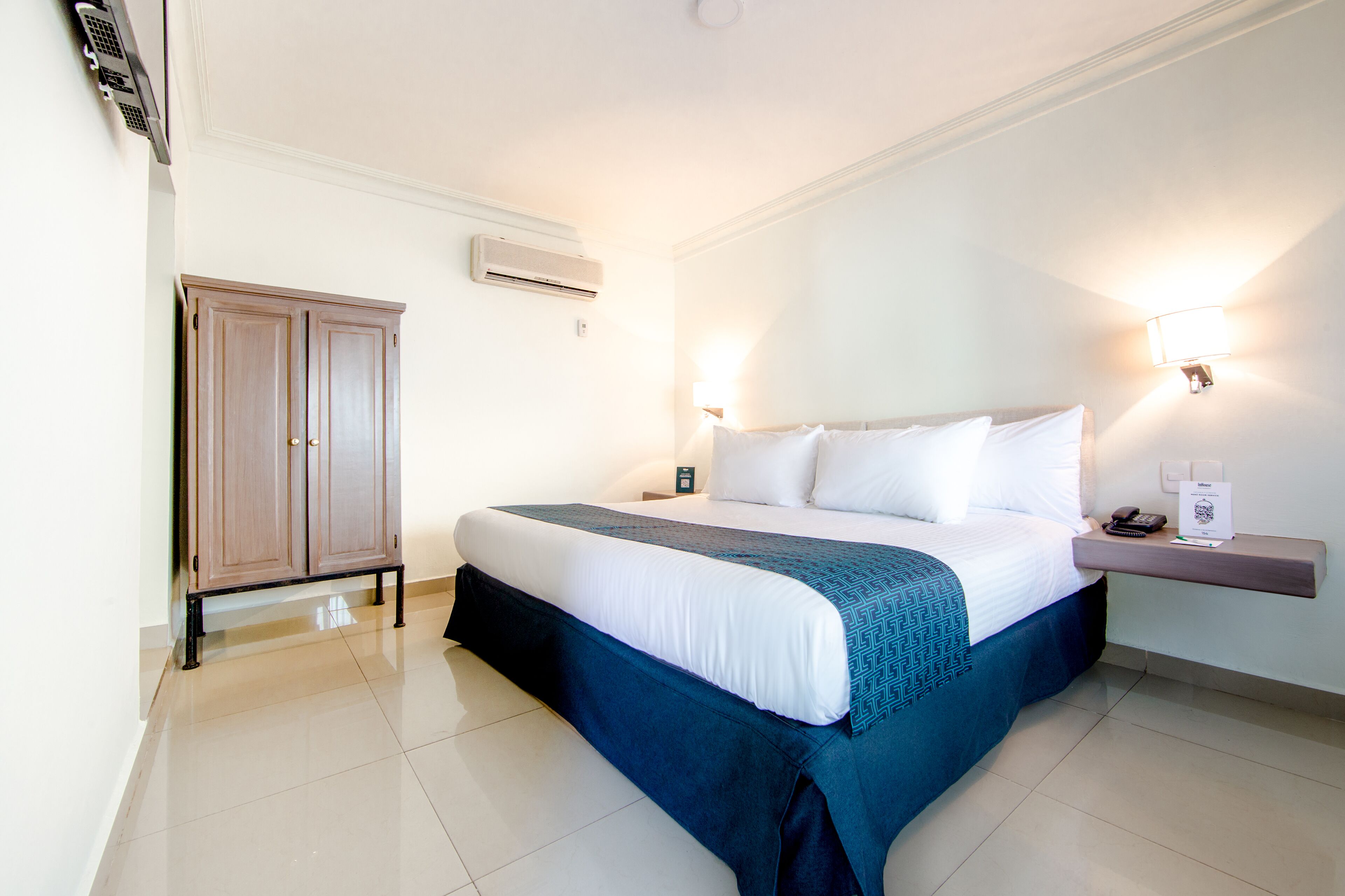 A hotel room with a large bed, a wardrobe, a bedside table with a lamp, and an air conditioning unit on the wall.