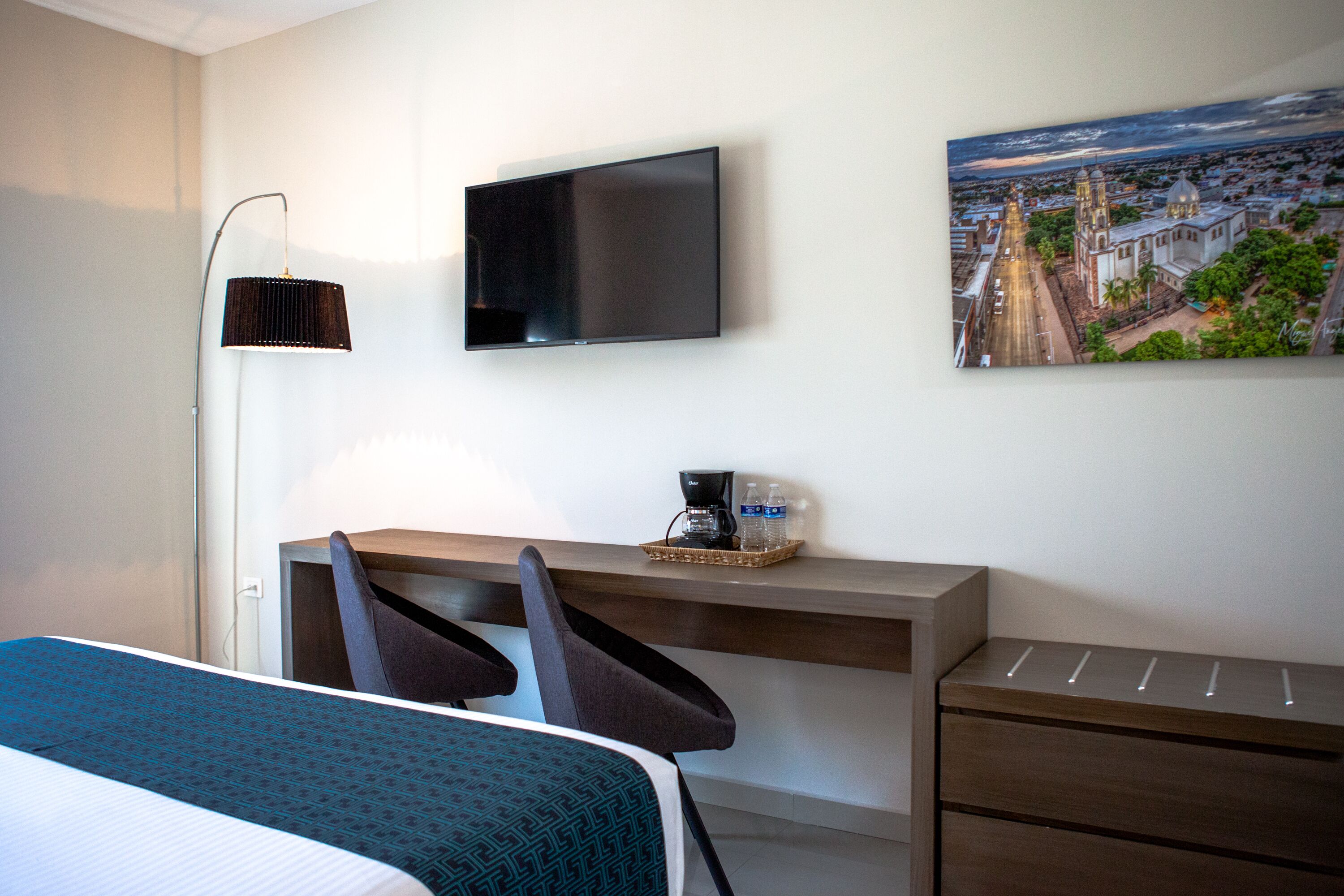 Habitación de hotel con cama, escritorio, sillas, televisor de pantalla plana, lámpara, cafetera, botellas de agua y un cuadro de un paisaje urbano.