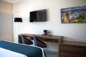 Habitación de hotel con cama, escritorio, sillas, televisor de pantalla plana, lámpara, cafetera, botellas de agua y un cuadro de un paisaje urbano.
