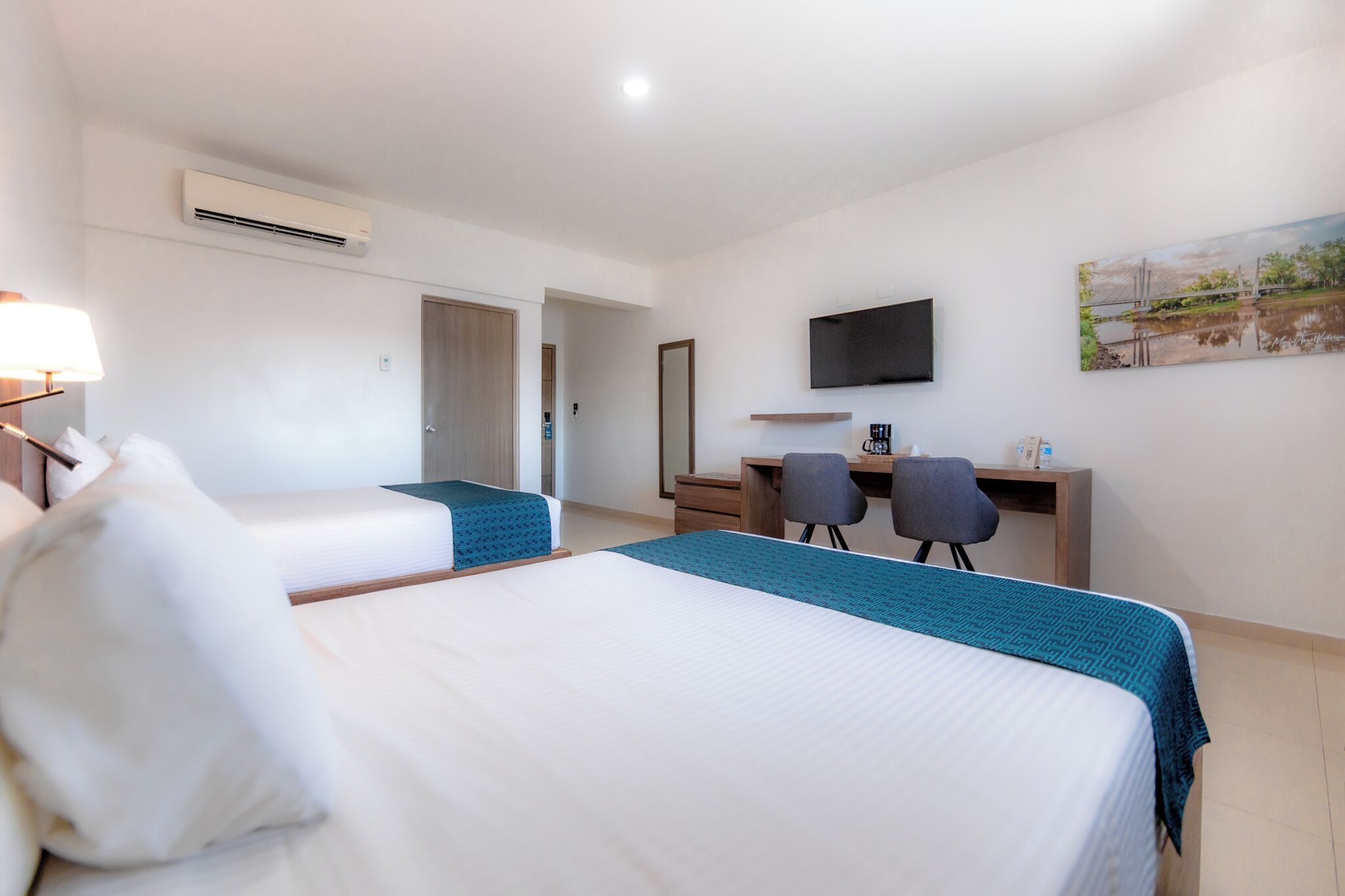 Habitación de hotel con dos camas, un escritorio con silla, un televisor montado en la pared y un equipo de aire acondicionado.
