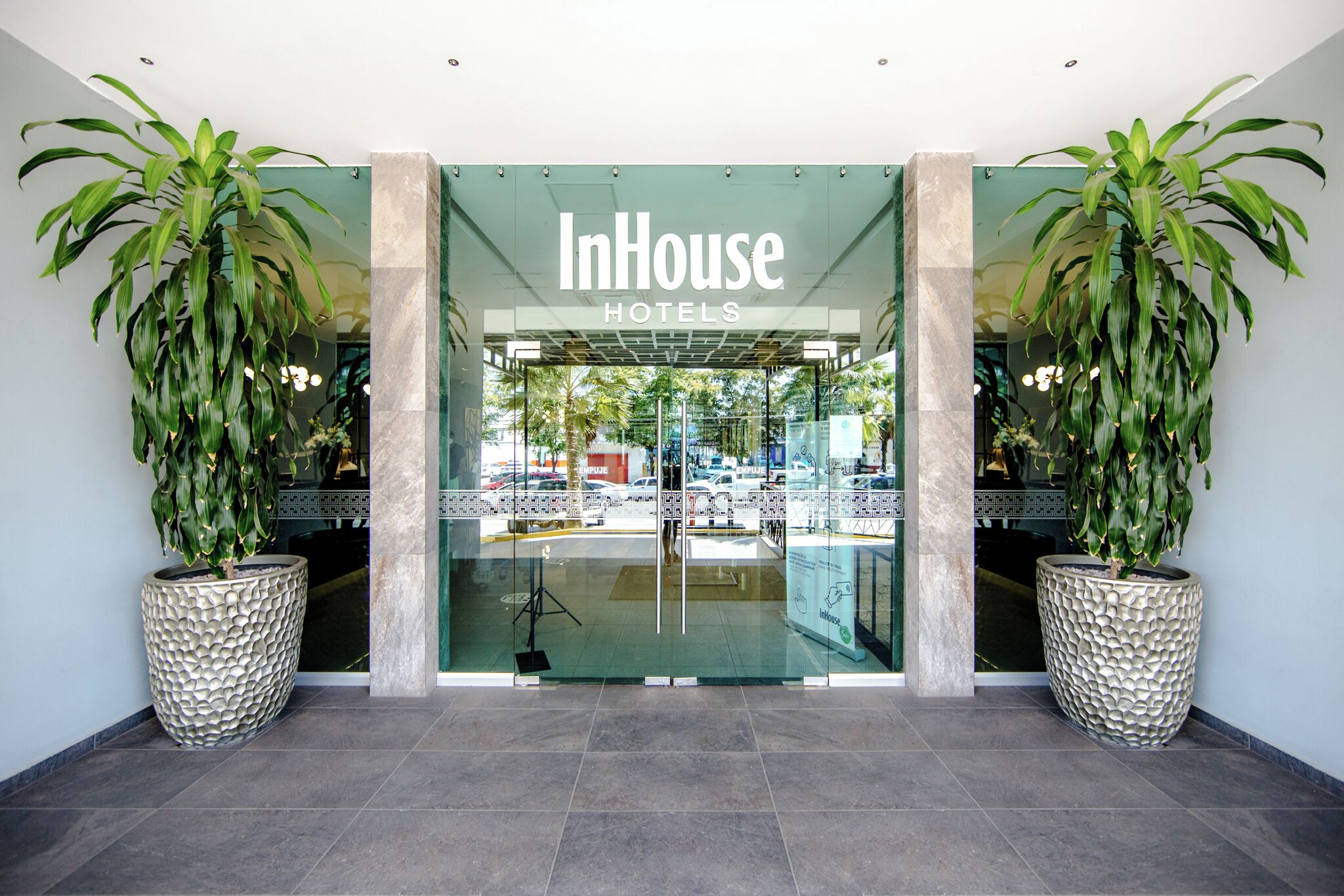 The entrance of InHouse Hotels with a glass door, two large potted plants, and a tiled floor.