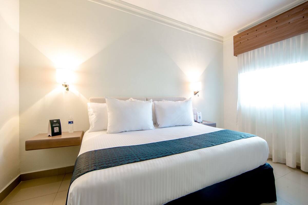 Habitación de hotel con cama, almohadas, mesita de noche y lámpara de pared, además de ventana con cortinas.