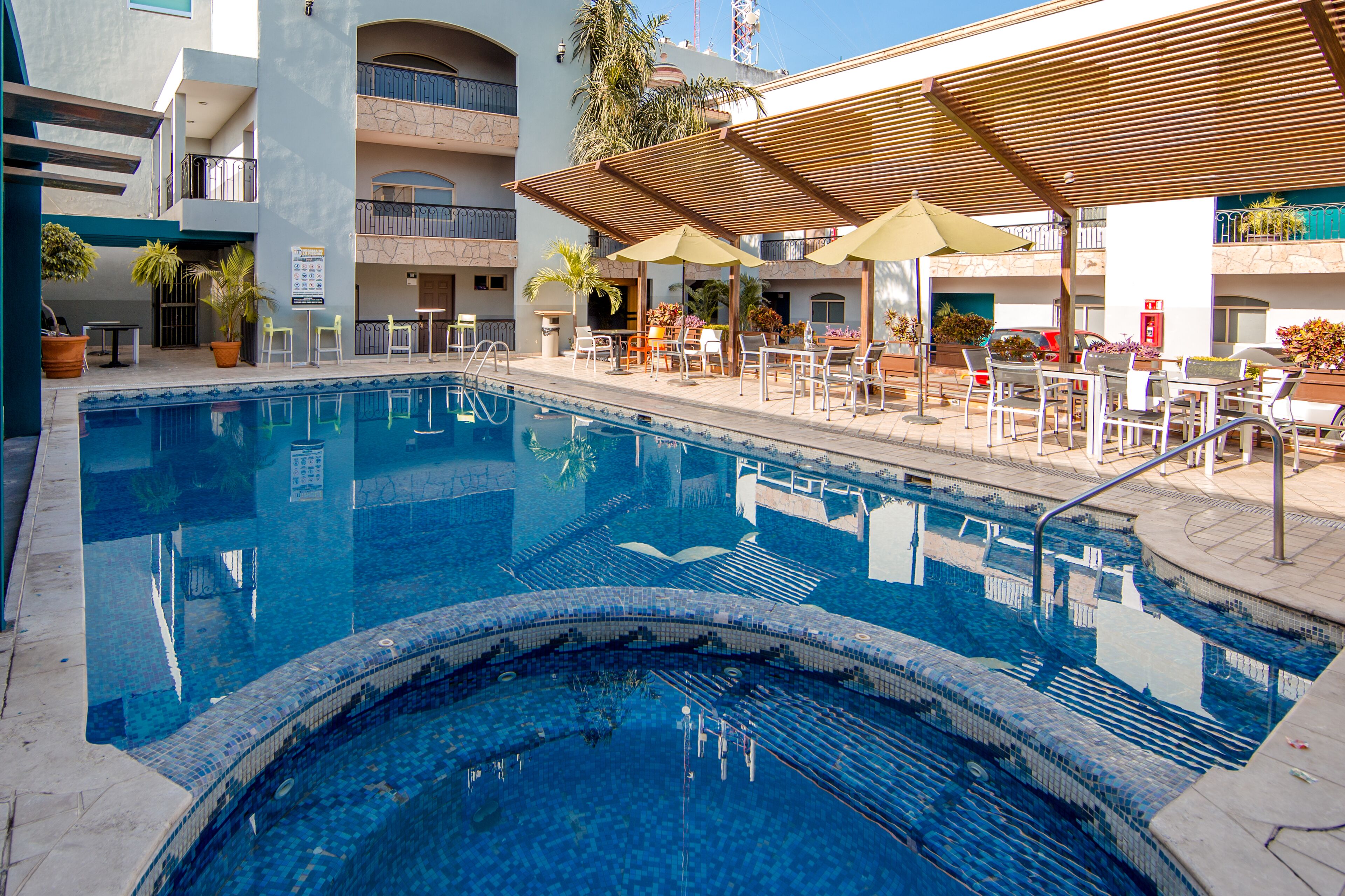 A swimming pool with lounge chairs and umbrellas, surrounded by a multi-story building with balconies.