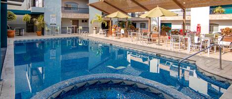 A swimming pool with lounge chairs and umbrellas, surrounded by a multi-story building with balconies.
