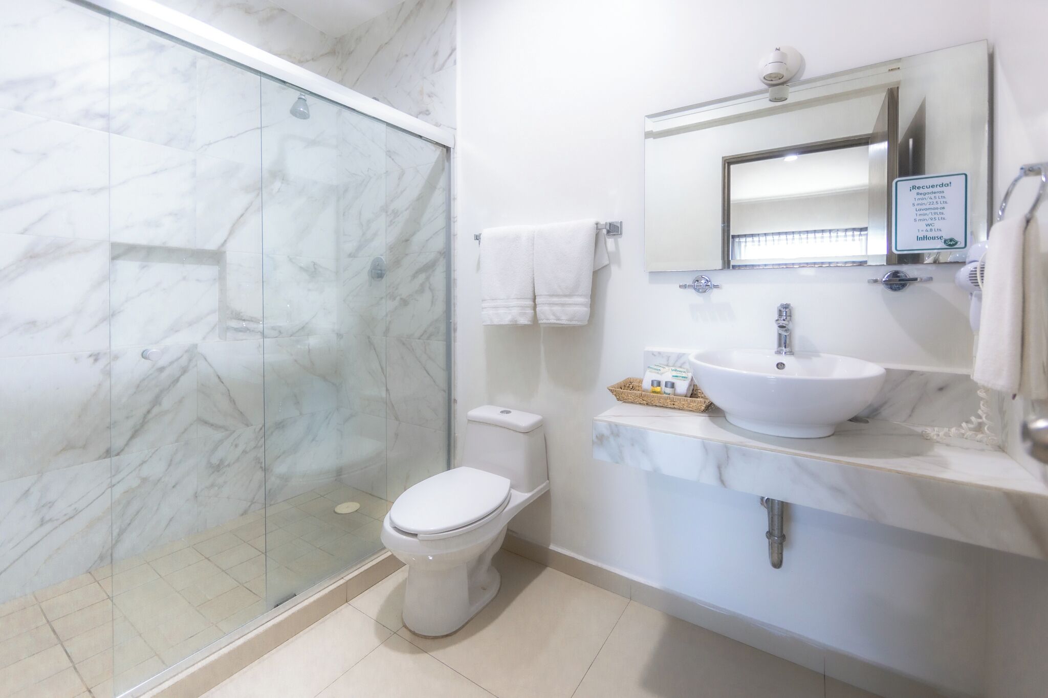A modern bathroom with a glass shower enclosure, a white toilet, a marble sink, and a mirror with a sign above it.