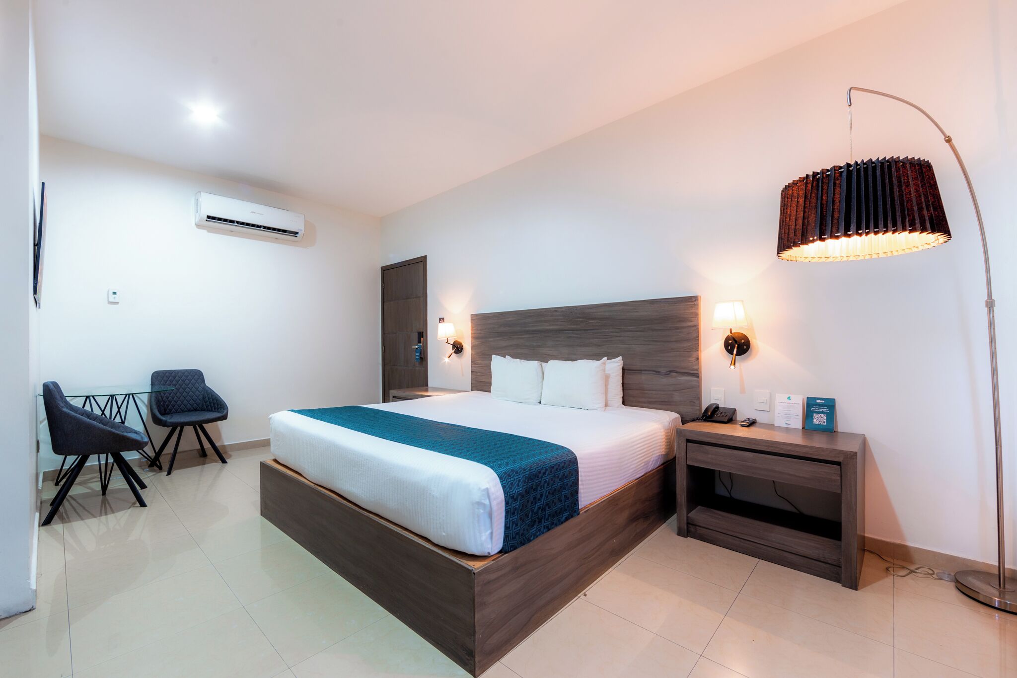 A hotel room with a bed, bedside table, lamp, chair, and an air conditioning unit.