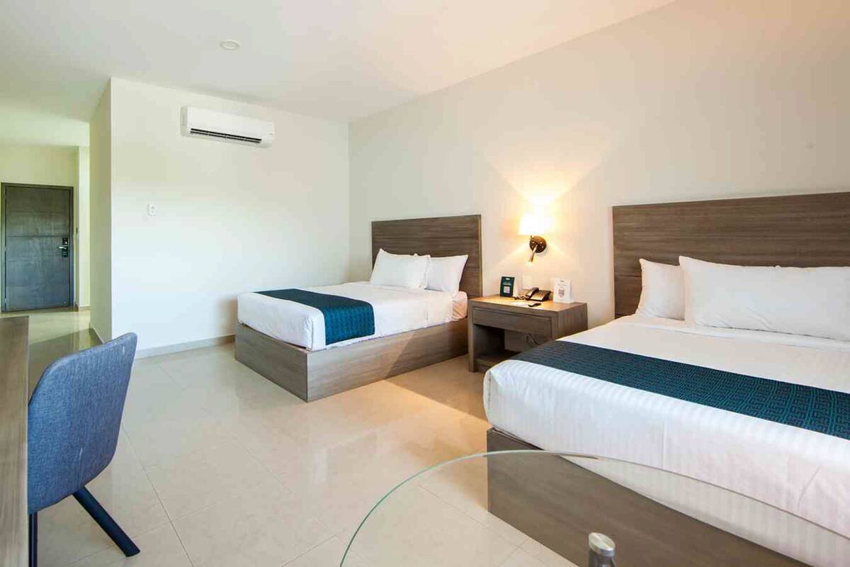 Habitación de hotel con dos camas, un escritorio, una silla, una mesita de noche con lámpara y un equipo de aire acondicionado.