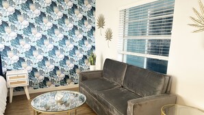 A living room with a grey sofa, a glass coffee table, a white side table, a patterned wallpaper, and a window with blinds. - Sunburst Inn (Indian Shores)