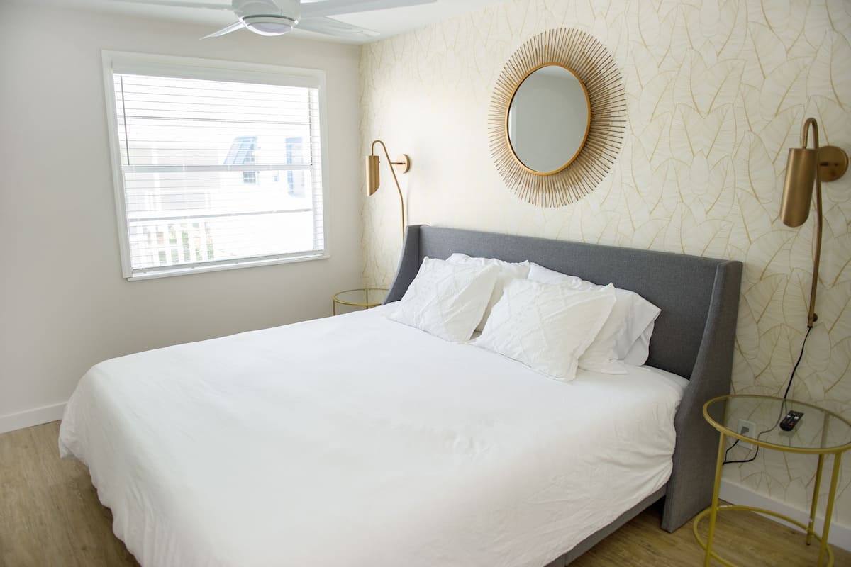 A neatly made bed with white linens, a round mirror with a gold frame, and a modern floor lamp.